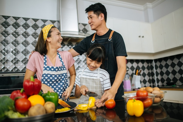 Aiuto sveglio della ragazza i suoi genitori stanno tagliando le verdure e stanno sorridendo mentre cucinano insieme nella cucina