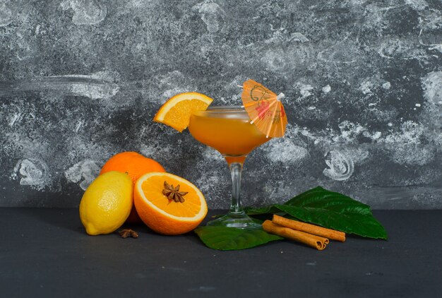 Alcune arance con succo in un bicchiere, limone, foglie su superficie scura e strutturata, vista laterale.