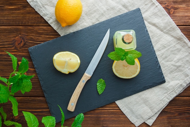 Alcune fette di limone con cartone nero, bottiglia di succo, coltello di legno in una ciotola su una superficie di legno, vista dall'alto.