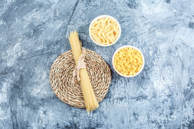 Alcuni assortiti di pasta in ciotole su intonaco grigio e sfondo tovaglietta di vimini, vista dall'alto.