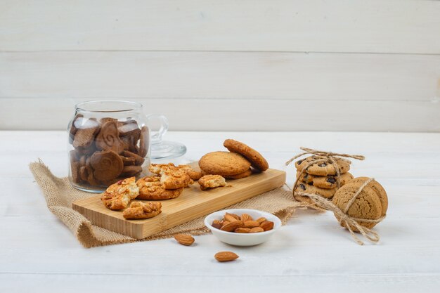 Alcuni biscotti marroni con mandorle in una ciotola, biscotti su un tagliere e un pezzo di sacco in un vasetto di vetro sulla superficie bianca