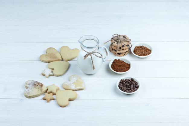 Alcuni diversi tipi di biscotti con chicchi di caffè, caffè istantaneo, cacao, brocca di latte sul fondo del bordo di legno bianco, vista ad alto angolo.