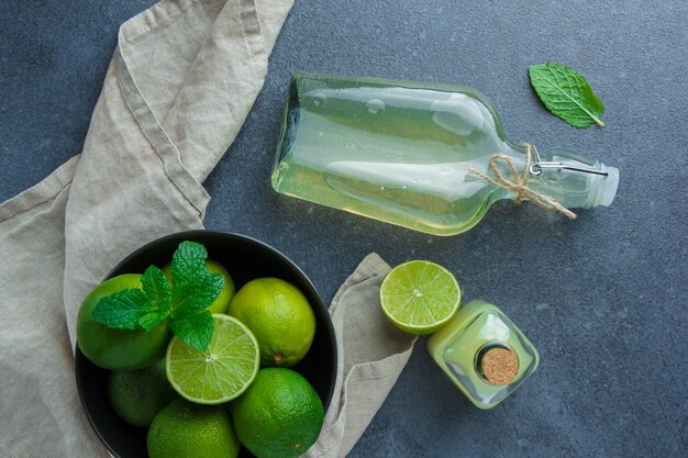 Alcuni limoni verdi con succo di limone in una ciotola nera su un panno di tessuto bianco su una superficie scura, distesi.