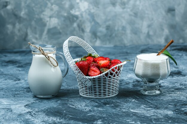 Alcuni un cesto di fragole con una brocca di latte e una ciotola di vetro di yogurt su sfondo di marmo blu scuro, primo piano.