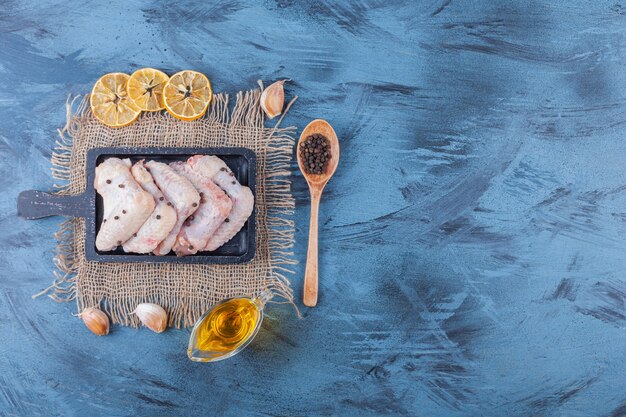 Ali di pollo su una tavola su un tovagliolo di tela accanto alla ciotola di olio, spezie, cucchiaio e limone essiccato sulla superficie blu