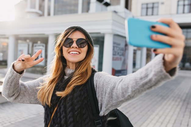 Alla moda affascinante bella donna in occhiali da sole moderni, caldo maglione invernale che fa il ritratto di selfie sulla strada nel centro della città. Look elegante, divertimento, espressione di emozioni positive e luminose.