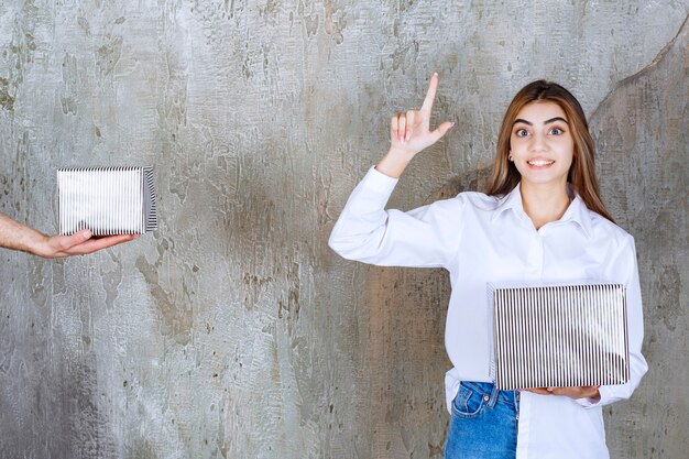 Alla ragazza in camicia bianca in piedi su un muro di cemento viene offerta una confezione regalo d'argento e ha una buona idea.