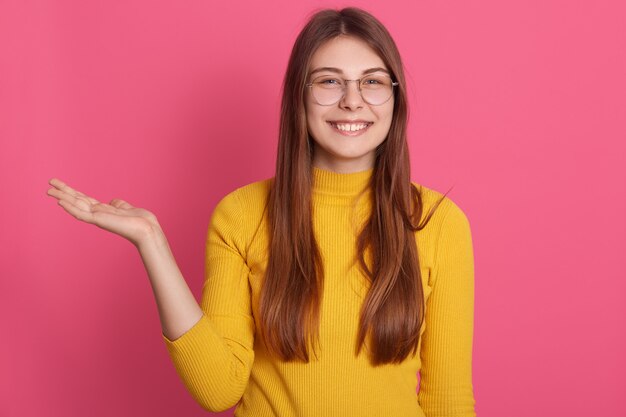 Allegra attraente giovane donna affascinante con divario nei denti, guardando direttamente alzando una mano, indossando la felpa gialla