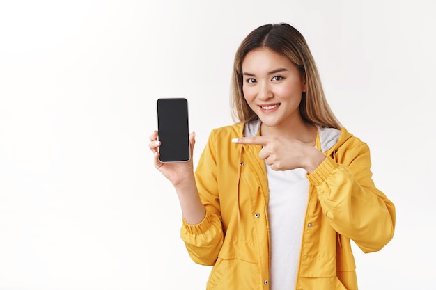 Allegra carina tenera donna bionda asiatica indossare giacca elegante gialla tenere smartphone mostra display puntare il dito indice schermo del telefono sorridente consigliare ampiamente app cool in piedi muro bianco