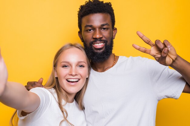 Allegra coppia interrazziale che scatta insieme un autoritratto, guardando davanti e sorridendo, posando su un muro giallo