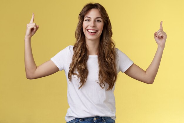 Allegra divertita felice vivace giovane ragazza taglio di capelli ricci lunghi alzare le mani rivolte verso l'alto sorridente a trentadue denti allegramente macchina fotografica presentare ottima variante suggerire di cliccare sul link del sito pubblicità sfondo giallo