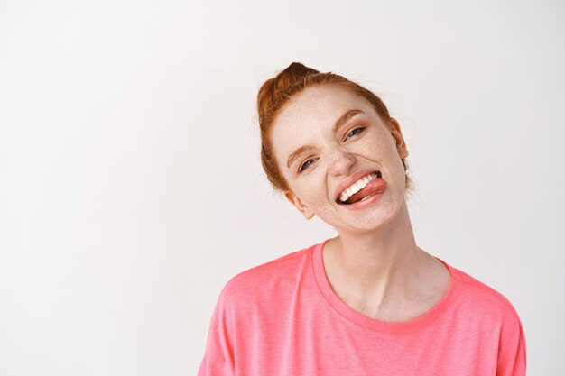 Allegra donna adolescente con i capelli allo zenzero pettinati in un panino disordinato, mostrando un sorriso bianco e la lingua, in piedi contro il muro bianco in t-shirt rosa