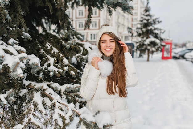 Allegra donna bianca indossa cappello lavorato a maglia e cappotto caldo in posa con un sorriso gentile accanto all'albero. Modello femminile estatico con capelli lunghi in giacca con pelliccia godendo le vacanze invernali all'aperto.