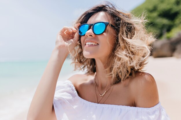 Allegra donna bionda in occhiali da sole guardando il cielo. Ritratto all'aperto di incantevole donna caucasica agghiacciante alla spiaggia sabbiosa.