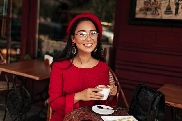 Allegra donna bruna dai capelli lunghi in berretto luminoso e vestito rosso sorride ampiamente e si siede in un bellissimo caffè La giovane signora asiatica con gli occhiali tiene una tazza di caffè bianca