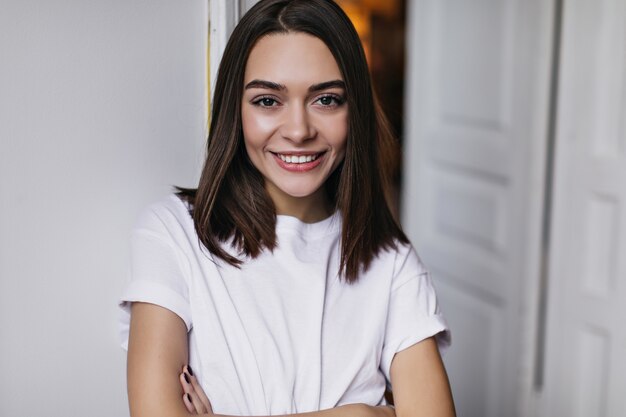 Allegra donna caucasica in maglietta bianca sorridente mentre si raffredda nel suo appartamento. Ritratto dell'interno del modello femminile felice con capelli castano scuro lisci.