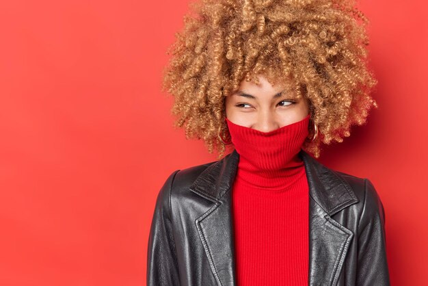 Allegra donna dai capelli ricci distoglie lo sguardo felicemente indossa dolcevita copre bocca con colletto vestito con giacca di pelle nera isolato su sfondo rosso vivido spazio vuoto per la tua pubblicità