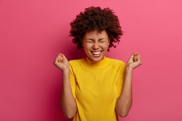 Allegra donna dai capelli ricci stringe i pugni con espressione positiva, si rallegra di notizie meravigliose, sorride felicemente, socchiude gli occhi con piacere, indossa abiti gialli, si erge contro il muro vibrante rosa