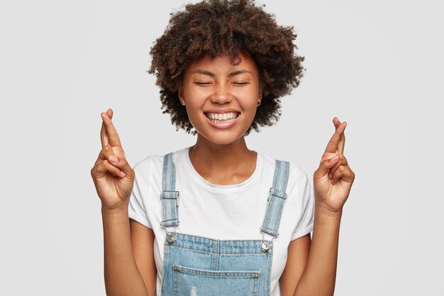 Allegra donna di colore con un sorriso a trentadue denti trattiene il respiro dalla tentazione, prega che i sogni diventino realtà, alza le dita incrociate