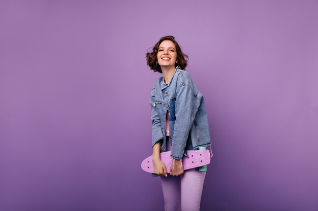 Allegra donna europea in pantaloni viola in posa con lo skateboard. Tiro al coperto di attraente ragazza sorridente con i capelli ondulati scuri.