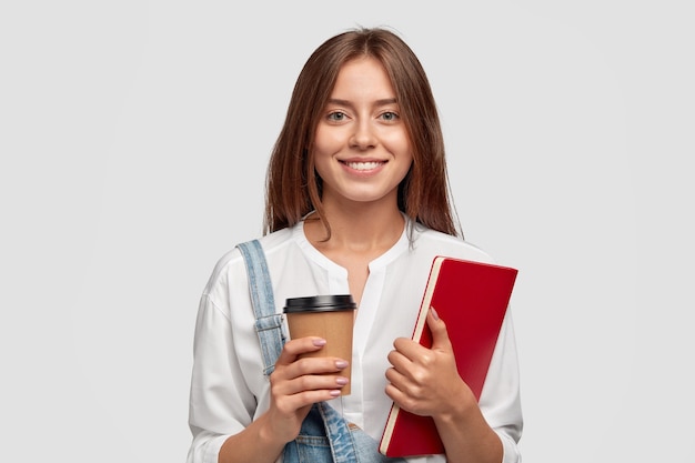 Allegra donna felice con un sorriso a trentadue denti, porta caffè da asporto e libro rosso, felice di finire lo studio