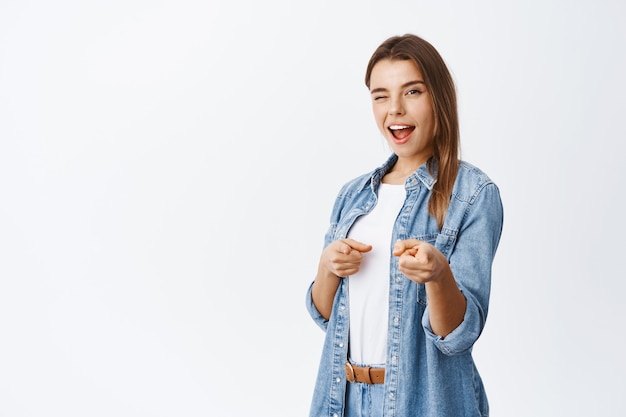 Allegra donna sorridente che invita o recluta, puntando il dito davanti e strizzando l'occhio per lodare qualcuno, in piedi in abito casual contro il muro bianco