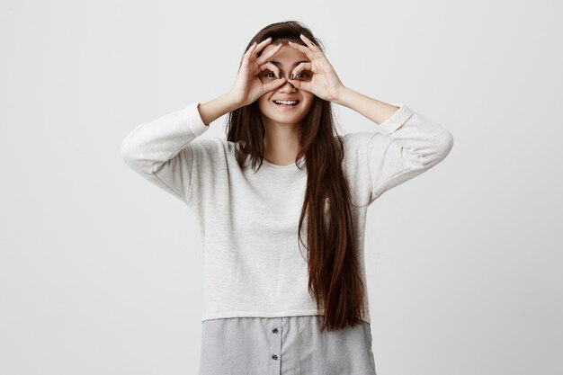 Allegra femmina adolescente eccitata con lunghi capelli lisci scuri che mostra gesti ok con entrambe le mani, fingendo di indossare gli occhiali e sorridendo ampiamente, godendosi la sua spensierata vita felice.