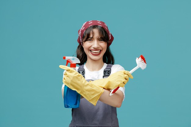 Allegra giovane donna addetta alle pulizie che indossa bandana uniforme e guanti di gomma che tengono spazzola e detergente incrociati guardando la fotocamera ridendo isolata su sfondo blu