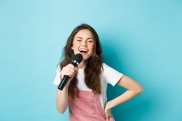 Allegra giovane donna che canta al karaoke, tenendo il microfono e sorridendo, divertendosi, in piedi su sfondo blu.