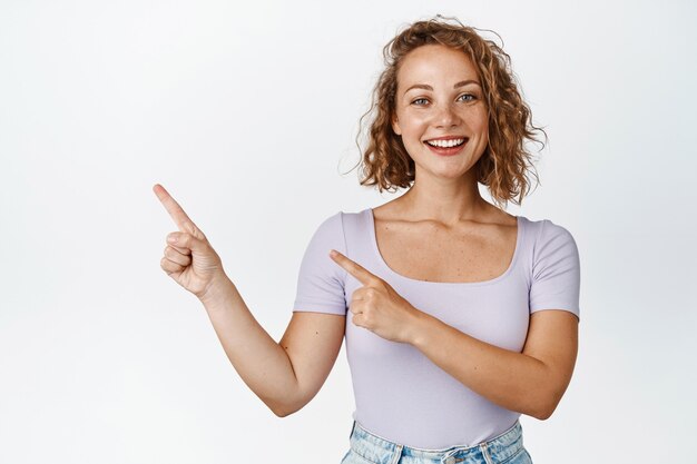 Allegra giovane ragazza bionda che mostra annuncio, puntando le dita a sinistra nello spazio della copia, logo aziendale, sorridente, in piedi su bianco