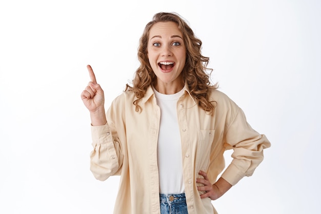 Allegra giovane ragazza bionda con i capelli ricci, vestiti alla moda, dito puntato nell'angolo in alto a sinistra, che mostra l'offerta promozionale, in piedi sul muro bianco