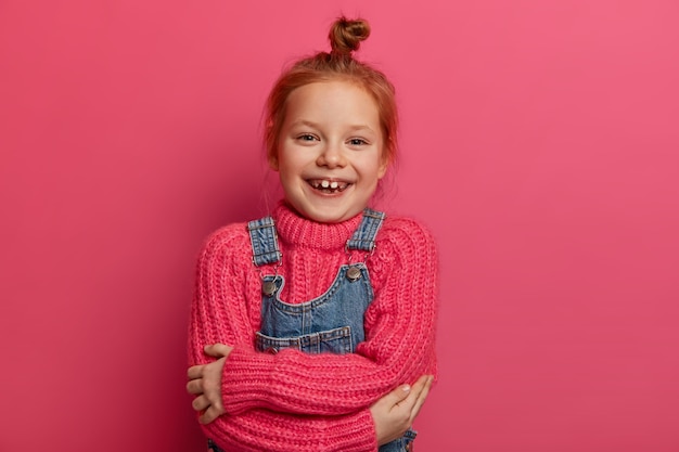 Allegra piccola ragazza rossa si abbraccia, si sente a suo agio, ha un nuovo maglione di lana rosa, un vestito morbido e caldo, sorride a trentadue denti, mostra i denti mancanti, ha i capelli rossi, isolato su un muro rosa.