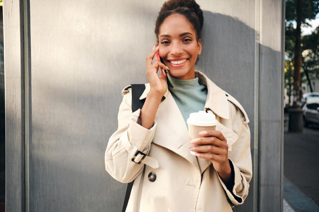 Allegra ragazza afroamericana in elegante trench con caffè per guardare con gioia nella fotocamera mentre si parla al cellulare all'aperto