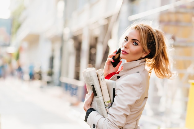 Allegra ragazza bionda con trucco luminoso che trasportano carta e laptop tornando a casa dall'ufficio