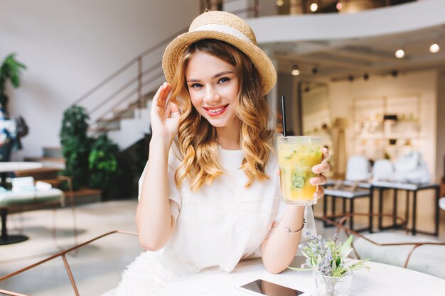 Allegra ragazza bionda con un bicchiere di cocktail di frutta rilassante nella caffetteria con interni moderni e sorridente dolcemente