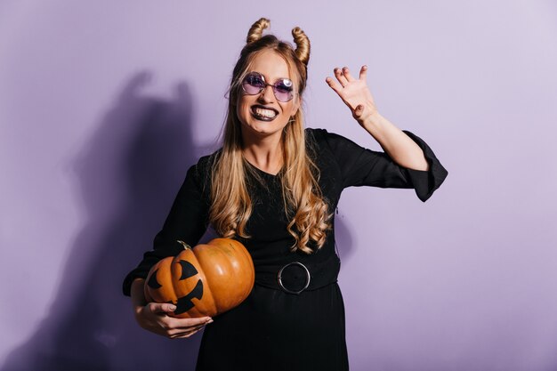 Allegra ragazza bionda in costume da strega godendo il carnevale. Tiro al coperto di sorridente signora spensierata con la zucca di Halloween in piedi sul muro viola.