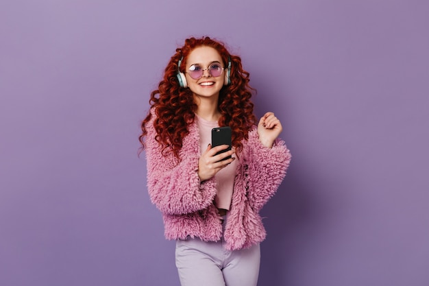 Allegra ragazza dai capelli rossi in eco-cappotto rosa e pantaloni leggeri sorridente. La donna in cuffie blu tiene lo smartphone nero.