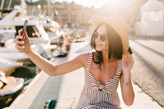 Allegra ragazza dai capelli scuri in occhiali da sole che fa selfie mentre riposa nel porto fluviale godendosi il sole. Ritratto all'aperto di giovane donna sorridente con cappello e vestito che si fotografa vicino a yacht.