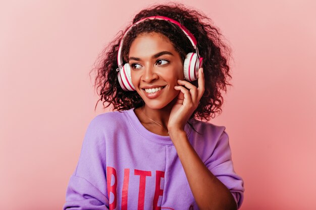 Allegra ragazza nera ascoltando musica pastello. Accattivante donna africana in maglione viola in posa in cuffia.