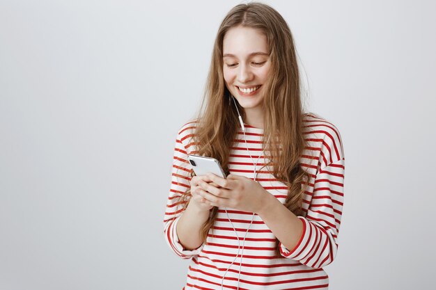 Allegra ragazza sorridente guardando lo schermo del telefono cellulare, indossando gli auricolari