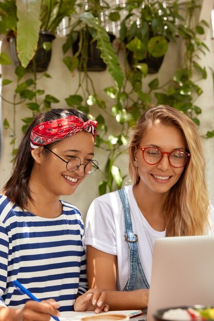 Allegre ragazze castane e bionde utilizzano un moderno dispositivo portatile per l'intrattenimento e lo studio, trascorrono il tempo libero insieme