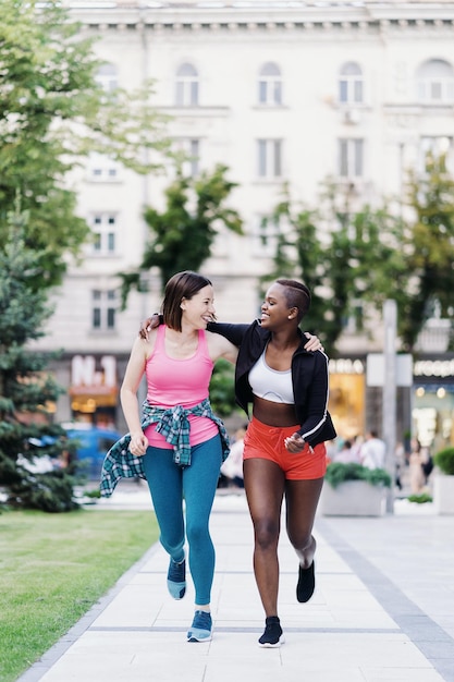 Allegri amici sorridenti in abbigliamento sportivo che corrono in città a discutere di donne multietniche che fanno un allenamento fitness