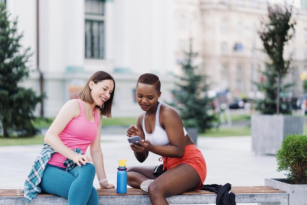 Allegri amici sorridenti in abbigliamento sportivo seduti su una panchina in città che discutono mentre utilizzano lo smartphone cercando di schermare le donne multietniche che hanno una pausa di allenamento fitness