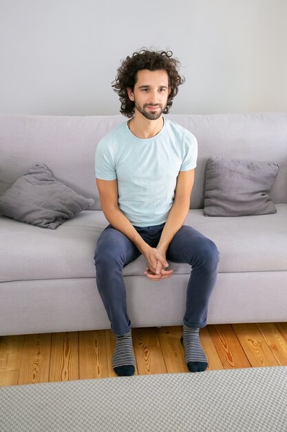 Allegro attraente giovane dai capelli ricci indossando t-shirt casual, seduto sul divano a casa, guardando lontano e sorridente. Colpo verticale. Concetto di ritratto maschile
