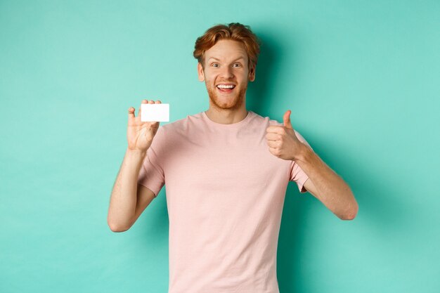 Allegro cliente di banca maschio in t-shirt che mostra il pollice in su e carta di credito in plastica, sorridendo soddisfatto alla telecamera, in piedi su sfondo turchese.