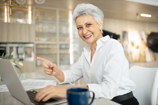 Allegro ed elegante donna matura manager che lavora in ufficio, seduto alla scrivania con generico computer portatile, sorridente ampiamente, puntando il dito anteriore, godendo il suo lavoro