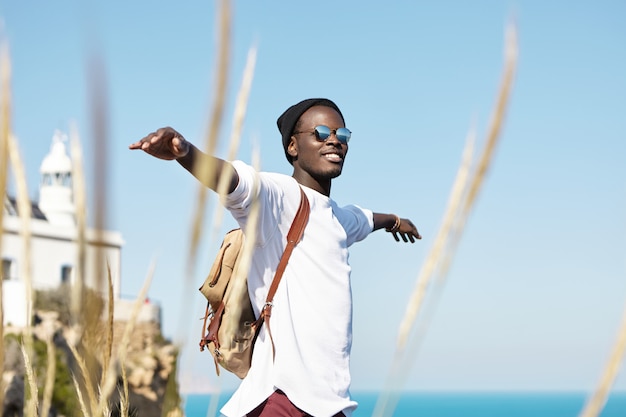 Allegro elegante giovane viaggiatore afroamericano con zaino che sorride con gioia, allargando le braccia, sentendosi libero, felice e rilassato, godendosi una bella giornata estiva mentre trascorri un weekend all'estero in riva al mare