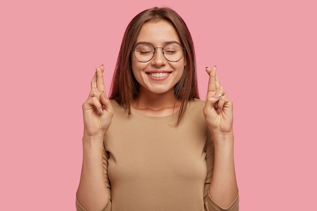 Allegro giovane femmina piuttosto giovane con espressione positiva incrocia le dita, spera di realizzare il sogno, esprime desideri, indossa occhiali rotondi e maglione casual, isolato su muro rosa.
