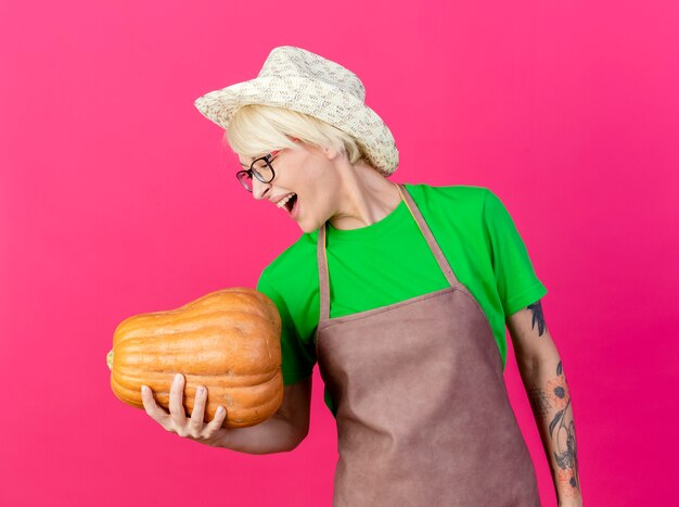 Allegro giovane giardiniere donna con i capelli corti in grembiule e cappello tenendo la zucca