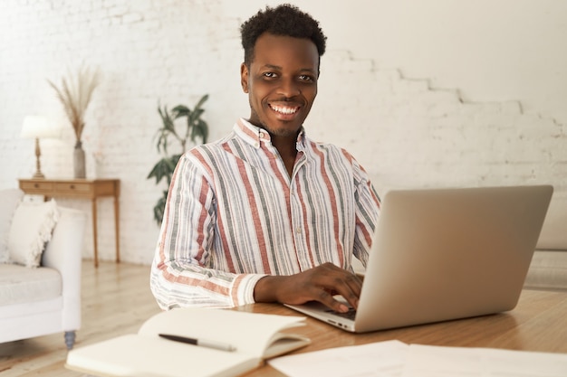 Allegro giovane studente africano seduto al tavolo in un accogliente soggiorno utilizzando il computer portatile per studiare tramite piattaforma online, prendendo appunti nel quaderno.